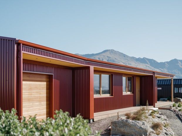 Single-storey contemporary residential building with a long, low-profile form, dark red corrugated metal cladding, timber garage door, recessed entry and windows, and a sheltered verandah-like overhang. The building sits in a rocky landscaped setting with rugged mountains visible behind it.