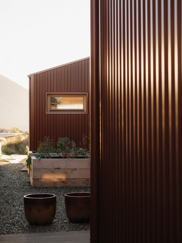 Exterior architectural view of a compact modern building clad in dark reddish-brown vertical corrugated metal, with a narrow horizontal window, rainwater downpipe, gravel courtyard, raised timber planter, and dark planters. The warm low-angle light and corrugated facade create a refined, minimalist composition.