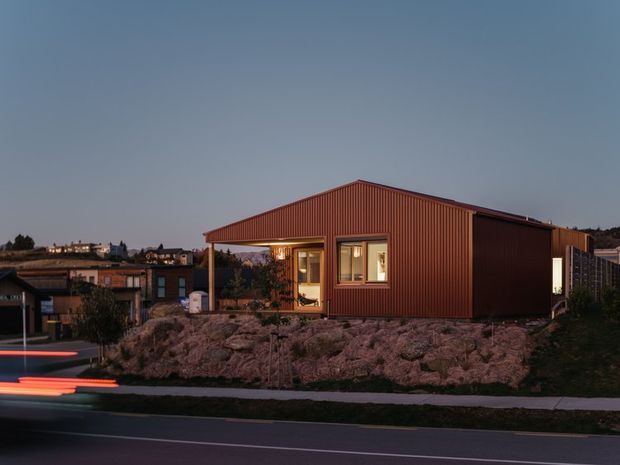 A compact single-storey house photographed at dusk, set on a rocky landscaped site above a street. The building has a simple gabled form with vertically ribbed reddish-brown metal cladding, a covered front porch, warm-lit rectangular windows, and a dark side wall. Motion-blurred vehicle lights appear on the road in the foreground, with neighbouring homes visible in the background.