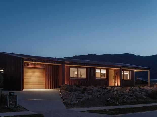 Twilight exterior view of a contemporary single-storey home on a sloping, rocky site. The long low building has dark vertical cladding, a shallow pitched roof, warm interior lighting, a timber-toned garage door, and several windows facing the street. A paved driveway and walkway lead to the house, with a dark mountain ridge visible in the background.