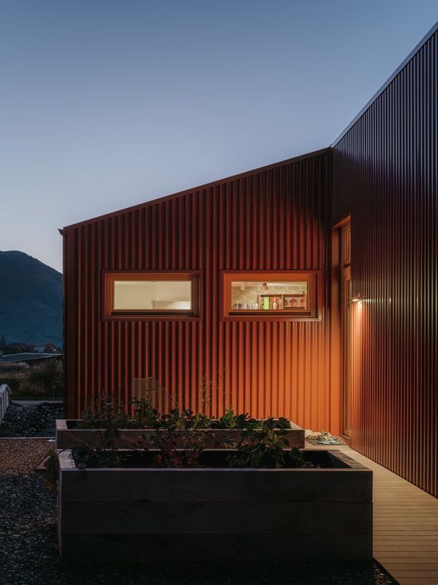 A dusk exterior view of a compact contemporary building clad in dark vertical corrugated metal, with warm interior light glowing through horizontal windows. A tall matching ribbed wall forms a narrow timber-decked passage beside raised garden planters. Mountainous terrain is visible in the background.