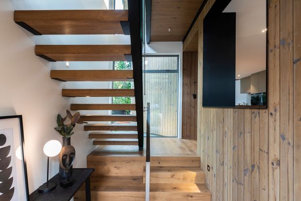 Interior view of a contemporary timber-lined stairway with open-riser wooden steps, a dark structural spine and slender glass balustrade. Vertical timber wall panelling continues beside the stairs, with a small landing and glazed door at the upper level. Black accents, a wall-mounted screen or artwork, console table and sculptural decor create a warm, refined residential entry circulation space.