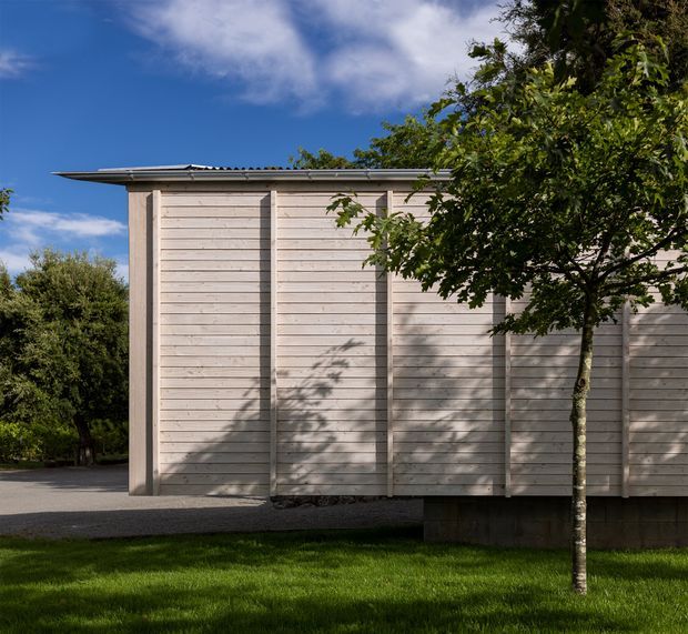 Exterior view of a simple single-storey structure clad in pale horizontal weatherboards, with prominent vertical battens, a shallow corrugated roof edge, and a dark concrete block base. A small tree and lawn occupy the foreground, with a paved path along the building and a clear blue sky behind.