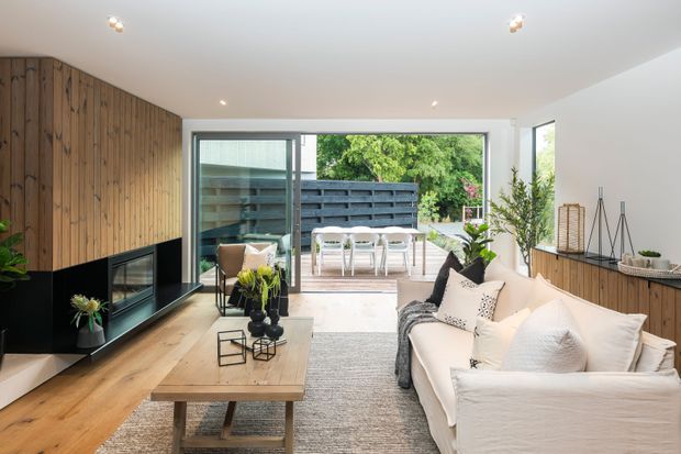 Bright contemporary living room with a warm timber feature wall and integrated black fireplace, pale timber flooring, a large light sofa, and a timber coffee table. Full-width sliding glass doors open directly to a furnished outdoor deck or patio, creating a strong indoor–outdoor connection. Built-in timber cabinetry runs along the right wall, with recessed ceiling lighting and neutral furnishings.