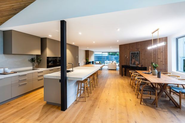 Open-plan contemporary kitchen, dining, and living area with a large central island, integrated cabinetry, timber flooring, and a warm timber feature wall. The kitchen has matte neutral cabinetry, a pale benchtop, and bar seating; the dining area includes a timber table beneath linear pendant lighting. A glazed opening at the rear connects to an outdoor area.