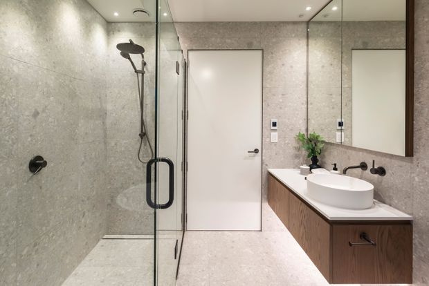 Contemporary bathroom with large-format grey stone-look wall and floor finishes, a frameless glass walk-in shower with black fixtures, and a floating dark timber vanity with a white benchtop and vessel basin. A large mirrored wall visually expands the space, which is clean and softly lit.