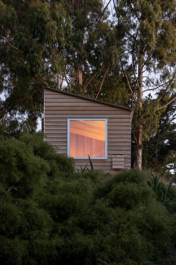 A small timber-clad structure nestled among dense eucalyptus and native vegetation. The simple facade has horizontal weatherboard cladding, a single sloping roof, and one large square window with a pale blue-grey frame. Warm timber interior surfaces are visible through the window, creating a softly illuminated focal point within the dark landscaped setting.