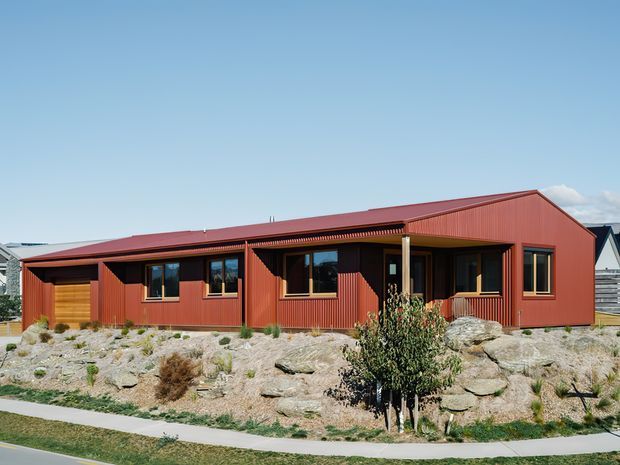 Single-storey contemporary residential house with a long low gabled form, deep covered veranda, and extensive red vertical metal cladding. The home sits on a landscaped rocky site with native-style planting and a concrete pedestrian path in front. Timber garage or entry door accents and dark-framed windows contrast with the red facade.