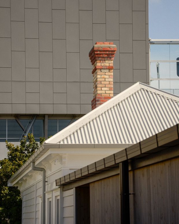Close-up architectural exterior showing a traditional white gabled roof and ornate eaves over a garage or outbuilding, with a red and cream brick chimney. A taller contemporary building with dark grey rectangular cladding and glazing forms the background, highlighting a juxtaposition of heritage detailing and modern construction.