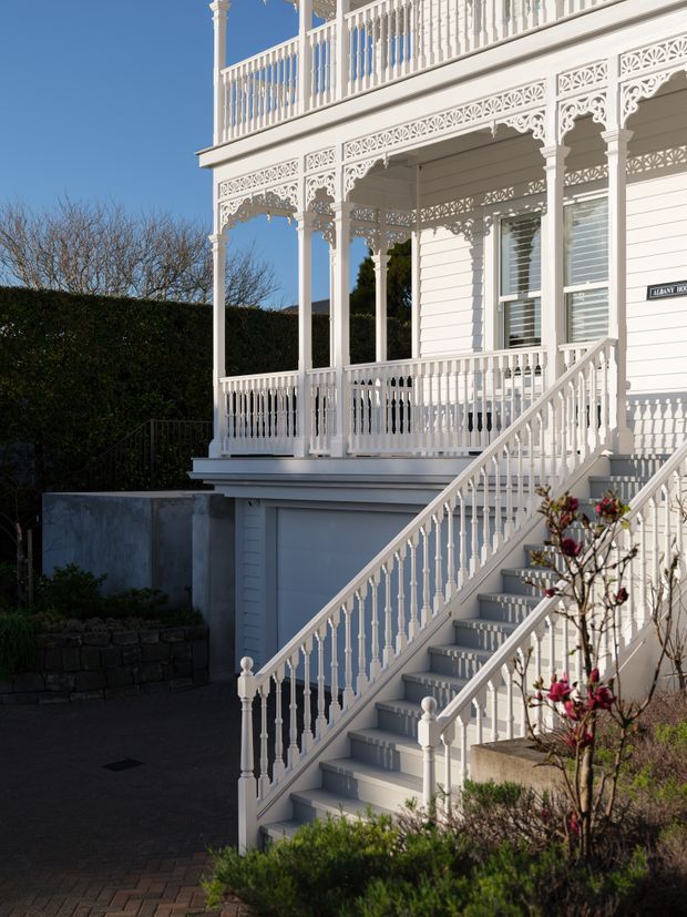 Exterior view of a highly detailed white heritage-style house, featuring ornate fretwork, decorative brackets, balustrades, turned posts, weatherboard cladding, and a prominent staircase leading to a raised veranda. The facade is brightly lit against a clear blue sky, with hedges and landscaped planting around the entry.
