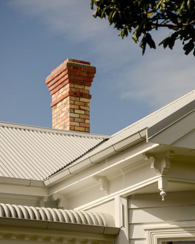 Close-up exterior architectural detail of a heritage-style white weatherboard building, showing ornate eaves brackets and cornice detailing, corrugated metal roofing, guttering, and a prominent exposed red-brick chimney against the sky.