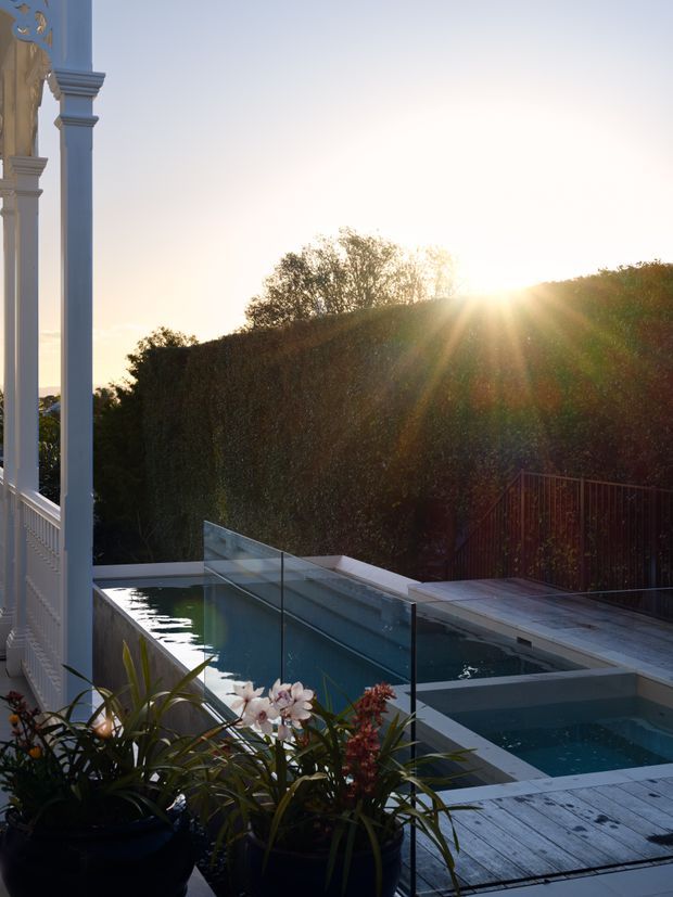 A sunlit outdoor pool terrace viewed beside a finely detailed white architectural veranda. The long rectangular pool and adjoining spa are edged with pale decking and enclosed by clear glass balustrades, with dense clipped hedging beyond. Potted flowering plants frame the foreground, creating a refined garden setting at sunset.