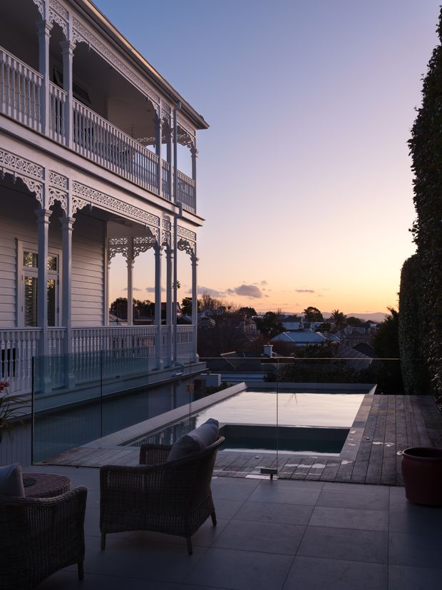 A heritage-style two-storey residence with ornate decorative ironwork verandahs overlooks a suburban landscape at sunset. In the foreground is a paved outdoor terrace with dark wicker seating, a glass balustrade, timber decking, and a compact rectangular swimming pool. The scene has a refined, tranquil atmosphere with warm dusk light.