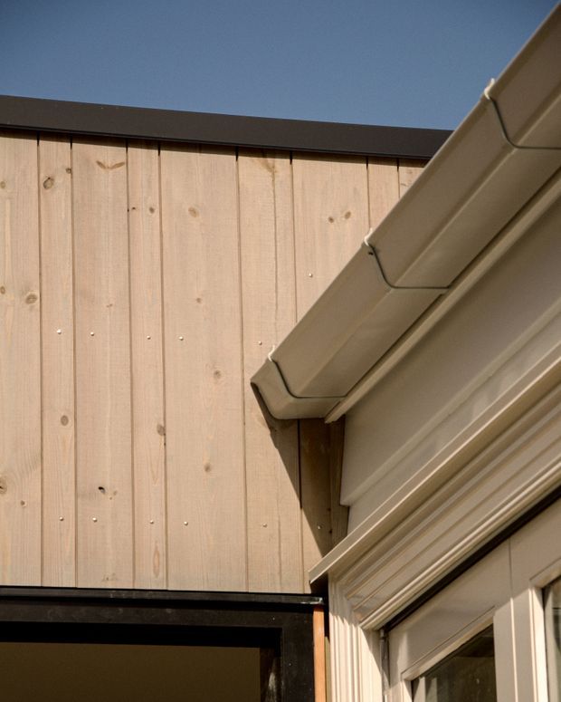 Close-up architectural detail showing a pale vertical timber-clad wall adjoining a traditionally detailed light-coloured building facade. The facade includes layered cornice and window or door trim, while a sloping eave with guttering projects diagonally across the upper right. Dark metal roof edges and a deep blue sky provide contrast.