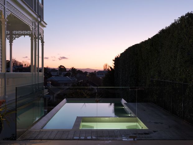Twilight exterior view of a refined residential pool terrace. A narrow rectangular swimming pool with an illuminated shallow section is bordered by timber decking, dark paving, and clear glass balustrades. At left is an ornate, elevated veranda with decorative metal fretwork and slender columns, while a tall clipped hedge screens the right side. Suburban rooftops and distant hills are visible beyond the terrace.