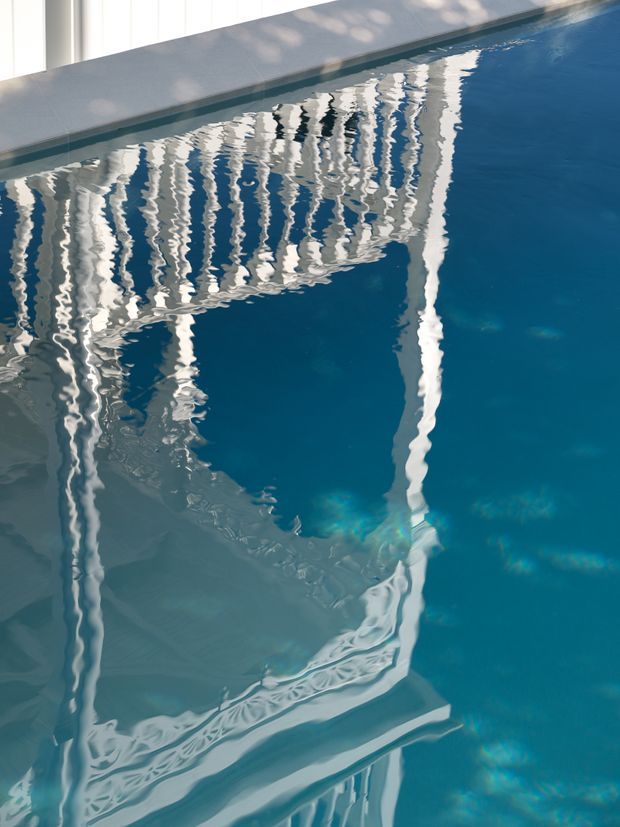 Close-up view of a clear blue swimming pool, with strong rippling reflections of white architectural glazing and structural elements across the water surface. The image is abstract and dominated by pool water and reflections, with a narrow pale pool edge visible at the top.
