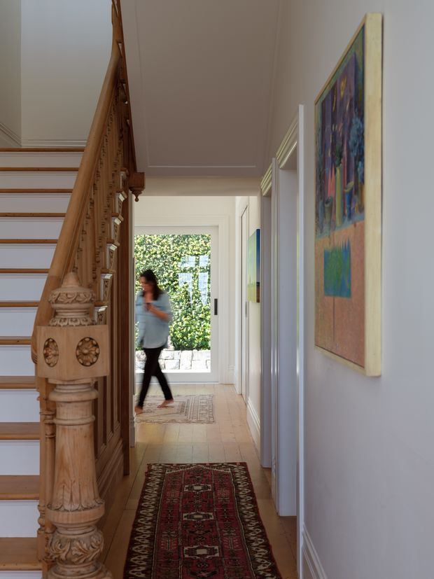 A long, refined residential hallway viewed beside an ornate timber staircase. The corridor has pale walls, detailed traditional stair balustrade and carved newel post, timber flooring, patterned runner rugs, framed artwork, and a glazed door at the far end opening toward a leafy exterior.