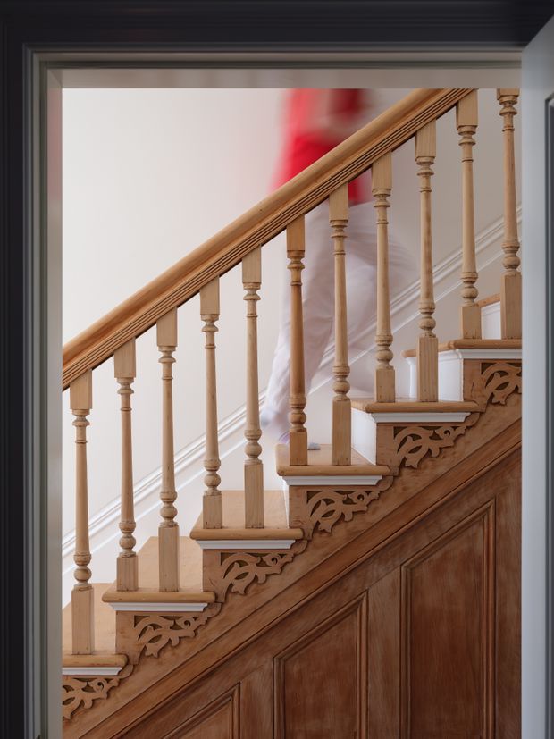 Close-up view of an ornate timber staircase with turned balusters, carved decorative brackets beneath the treads, a polished wooden handrail, and rich timber-panelled stair storage or wall lining. The staircase is framed by dark-painted trim, with a pale wall and a glimpse of red artwork or fabric in the background.