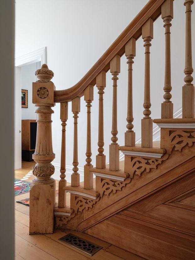 A close interior view of an ornate timber staircase and balustrade. The staircase features turned wooden spindles, carved decorative brackets and floral detailing, a substantial carved newel post, polished timber stair treads, and a timber floor with a decorative metal air vent. White walls and a doorway provide a light backdrop.