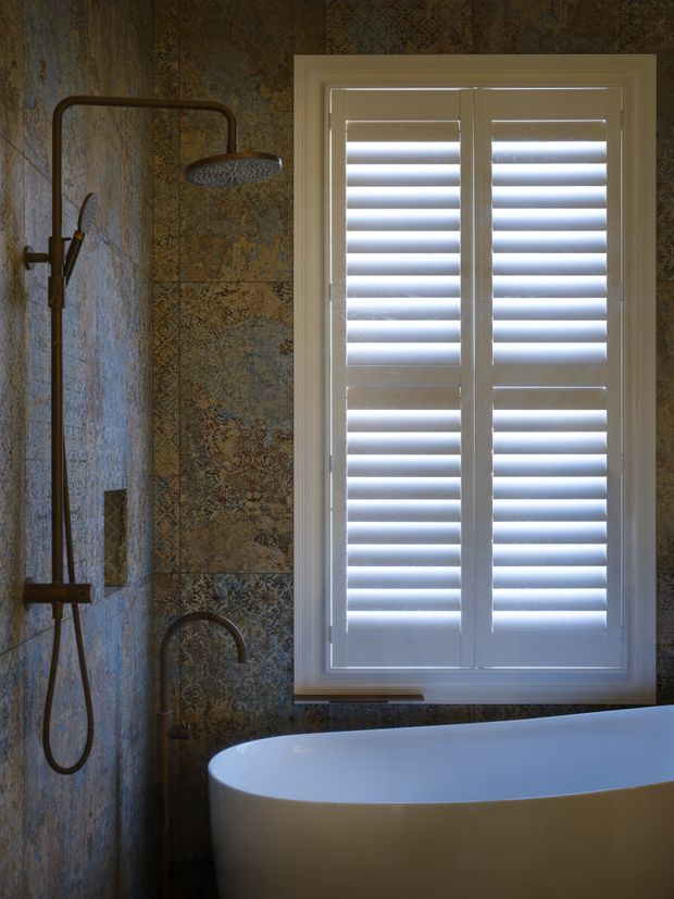 Moody bathroom interior featuring a freestanding white bathtub, dark bronze shower fittings, patterned decorative wall tiles, and a large white plantation-shuttered window. The low, atmospheric lighting creates a refined, traditional feel.