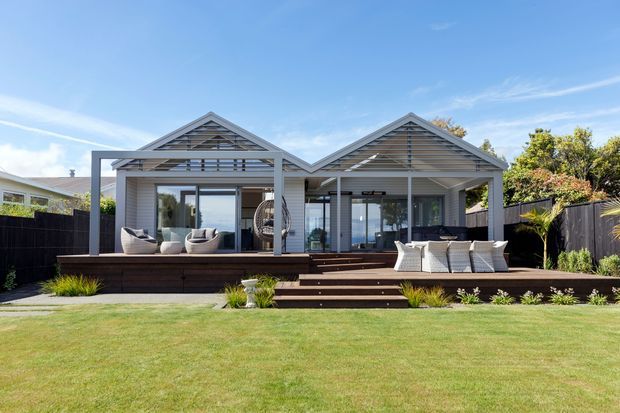 A single-storey contemporary coastal-style house viewed from the landscaped front lawn. The facade features two connected gabled roof forms, pale horizontal weatherboard cladding, dark-framed glazing, and a broad elevated timber deck with outdoor seating and steps leading to the lawn. The open deck and glazing suggest an outdoor living area directly connected to the interior.