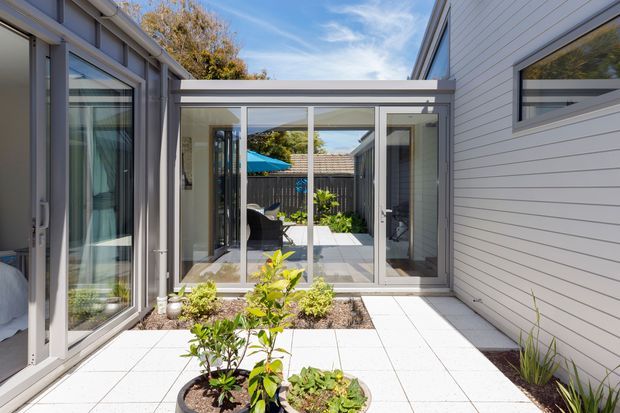 A bright enclosed courtyard between two contemporary residential wings, with pale tiled paving, low planted garden beds, extensive glazed doors and windows, and horizontal light-coloured cladding. The courtyard opens visually toward an outdoor seating area with a blue umbrella. The composition highlights the connection between indoor rooms and a landscaped central outdoor space.
