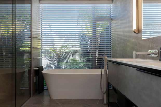 Contemporary bathroom featuring a freestanding white bathtub positioned in front of wide horizontal blinds, a glass-enclosed shower area, a floating vanity with a pale stone-look benchtop, and grey textured wall and floor finishes. Natural greenery is visible beyond the windows.
