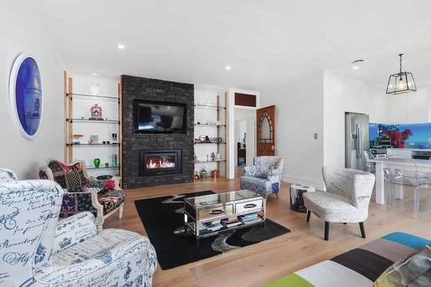 Bright open-plan living room with a dark brick fireplace wall, built-in shelving, television and firebox, surrounded by patterned upholstered armchairs and a glass-and-metal coffee table. A dining or kitchen area is visible beyond the living space, with timber flooring, white walls and a pendant light.