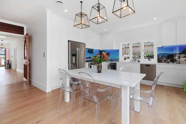 Bright, spacious contemporary kitchen and dining area with white full-height cabinetry, a large white island, stainless-steel appliances, pale timber flooring, transparent dining chairs, and three geometric pendant lights. The kitchen opens to a hallway through a decorative timber-framed opening, with coastal imagery visible on the splashback or screens.