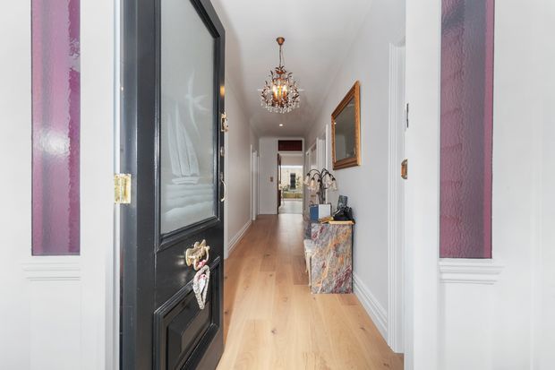 A bright, elegant residential hallway viewed through an open black front door. The long corridor has pale timber flooring, white walls with decorative moulding, purple inset wall panels, a crystal chandelier, framed artwork, and a marble-fronted console table with decorative objects. The space leads toward a glazed doorway at the far end.