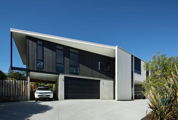 Exterior view of a contemporary two-storey residence with a sharply sloping mono-pitched roof, dark vertical timber cladding, light corrugated metal cladding, concrete garage surround, black-framed windows, and an open carport beneath the upper level. A broad concrete driveway and landscaped planting occupy the foreground.