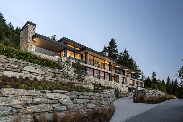 Exterior view of a contemporary multi-level hillside residence integrated into a steep, landscaped site. The house has long horizontal glazing, dark roof planes and framing, substantial natural stone walling, balconies, and a prominent stone-clad chimney or vertical tower. Terraced stone retaining walls and a curving driveway dominate the foreground, with dense mature trees surrounding the property.