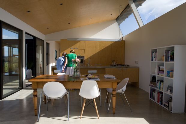 A bright, open-plan kitchen and dining area with full-height glazing and a high angled ceiling. The room features warm timber kitchen cabinetry and ceiling lining, a central island, a substantial timber dining table with modern white chairs, polished concrete flooring, and open white shelving. Strong sunlight enters through the glazing and clerestory-like roof windows, creating a spacious contemporary atmosphere.