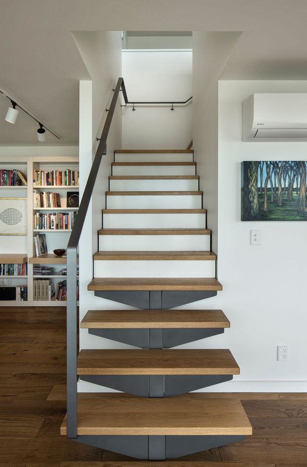 Interior view of a modern open-riser staircase with warm timber treads, dark grey steel stringer and balustrade, and white walls. The stair rises from a timber-floored living area with built-in bookshelves, track lighting, artwork, and a wall-mounted air-conditioning unit. A skylit or double-height stair void brightens the space.