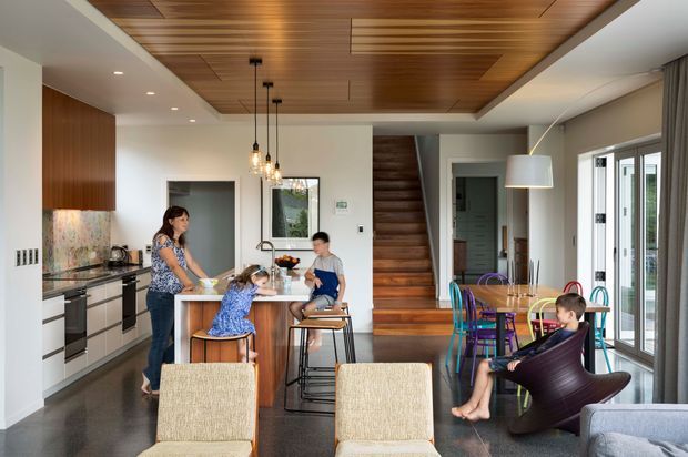 Open-plan contemporary kitchen, dining and living area with a timber-lined recessed ceiling, dark timber staircase, island bench, pendant lighting, and full-height glazed doors opening to the exterior. The kitchen has dark cabinetry, pale benchtops and a colourful patterned splashback. A family is gathered around the island and dining area.
