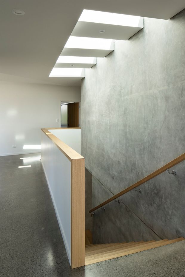 A bright contemporary stair landing with a stairwell descending beside a large textured grey feature wall. The space has polished concrete flooring, white plastered walls and ceiling, multiple rectangular skylights, and minimalist timber handrails and balustrade trim. A partially visible doorway opens beyond the landing.