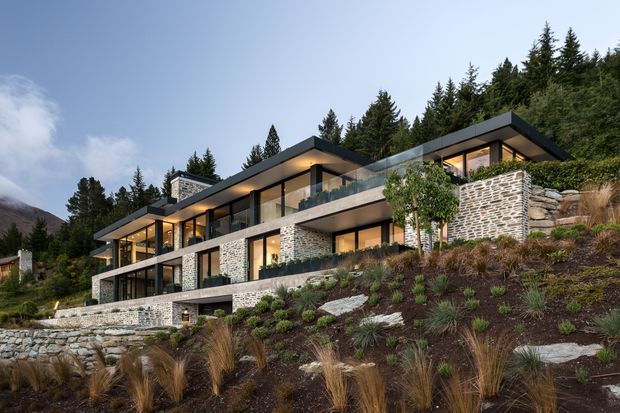 A large contemporary multi-level hillside residence set among dense evergreen trees and natural landscaping. The home features strong horizontal rooflines, extensive floor-to-ceiling glazing, projecting terraces with glass balustrades, dark metal framing, and extensive stacked natural-stone wall finishes. The sloped site is landscaped with native grasses, shrubs, boulders, and stone retaining walls, with mountain terrain visible in the background.