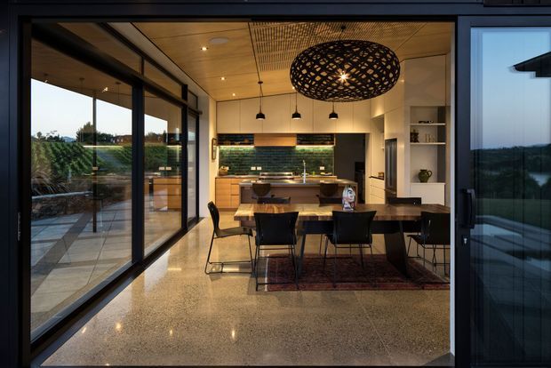 A contemporary open-plan kitchen and dining area viewed through large black-framed glazed doors. The space features a timber kitchen with island, dark green tiled splashback, polished terrazzo-like flooring, exposed timber ceiling detailing, extensive glazing, a dark timber dining table, black dining chairs, and a sculptural woven pendant light. A terrace and landscaped vineyard-like setting are visible beyond the glazing.