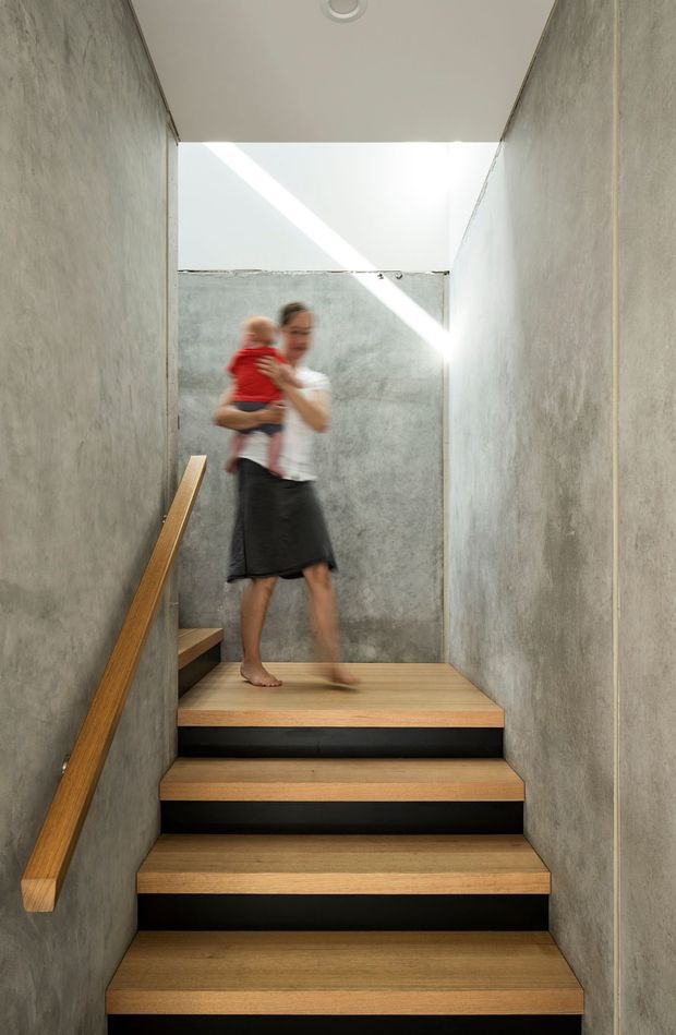 A narrow modern stairway with broad timber treads and dark contrasting risers, enclosed by raw grey concrete or cement-rendered walls. A simple timber handrail runs along the left wall. The stair rises toward a bright open skylit landing, with an adult carrying a child visible at the top. The composition emphasizes the textured concrete surfaces, warm timber, strong geometric lines, and natural daylight.