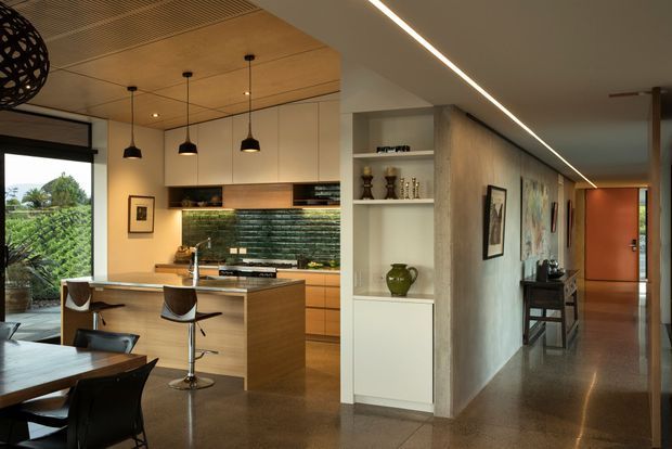 Open-plan contemporary kitchen and dining area opening into a long hallway. The kitchen features a timber island with two bar stools, integrated cabinetry, a dark textured tile backsplash, pendant lights, and a warm timber ceiling. A dining table occupies the foreground, while a built-in display niche and artwork line the hallway leading to a timber door. Large glazing provides views toward lush outdoor planting.