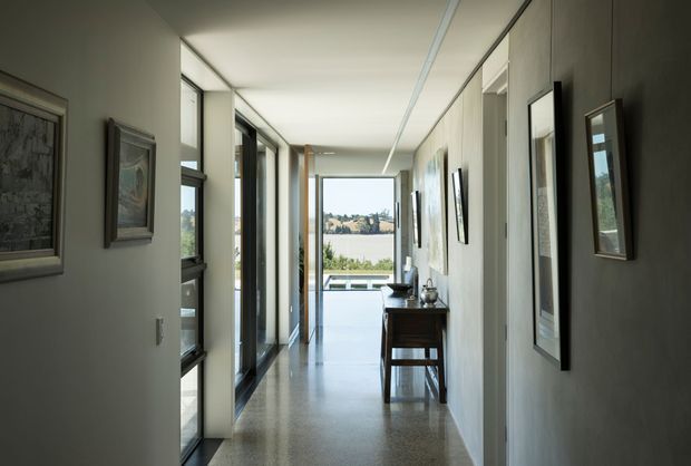 A long, bright residential hallway with polished concrete flooring, pale walls, extensive floor-to-ceiling glazing on the left, and framed artwork along both walls. A narrow timber console table sits midway down the corridor, which leads toward a glazed opening and a distant landscaped view. The space has clean contemporary detailing and strong linear perspective.