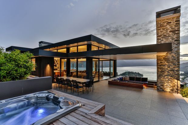 Contemporary multi-level flat-roof home with extensive floor-to-ceiling glazing, dark cladding, and a prominent stacked-stone chimney or feature wall. The image focuses on a spacious rooftop terrace overlooking a coastal or lakeside landscape, with an outdoor dining area, lounge seating, and a built-in spa set into timber decking. Warm interior lighting contrasts with the cool evening sky and broad water views.