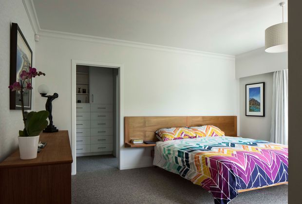 A bright, well-appointed bedroom with a built-in timber headboard and bedside shelf, colourful geometric-patterned bedding, grey carpet, white walls and ceiling, and a walk-in wardrobe visible through an open doorway. A timber dresser with an orchid and artwork sits to the left, while a pendant light and curtained window are visible on the right.