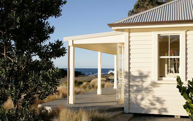 A coastal single-storey cottage exterior with cream horizontal weatherboard cladding, white trim, a covered open porch, and a corrugated metal hipped roof. The building overlooks a sandy shoreline and ocean, with native grasses and shrubs around a concrete path and terrace.