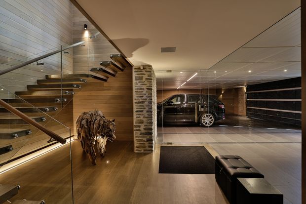 A sophisticated residential entry and garage interior viewed from a timber-floored foyer. The space features a floating timber staircase with clear glass balustrades and dark metal supports, a textured exposed-stone column, warm timber wall lining, recessed and linear lighting, and a large tiled garage beyond glazed partitions. A luxury vehicle is visible in the garage, with a black leather bench and dark entry mat in the foreground.