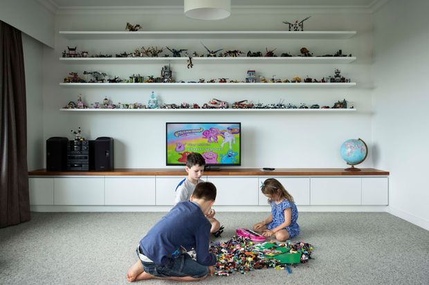 Bright, carefully designed children's play and media room with three children assembling toys on a large grey carpet. A long wall of white built-in cabinetry supports a television and stereo, while several floating display shelves hold extensive toy and model collections. The room has white walls, subtle ceiling moulding, a timber-toned benchtop, dark curtains, and a globe, creating a clean contemporary family space.