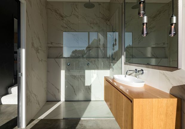 Bright contemporary bathroom with a walk-in glass shower, large marble-look wall and floor tiles, a floating timber vanity with a white vessel basin, wall-mounted chrome tapware, and a large mirrored cabinet. An open doorway connects directly to a carpeted bedroom or dressing area.