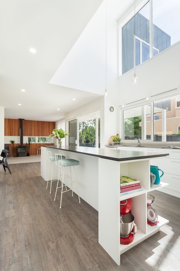 Bright, open-plan contemporary kitchen with a large white island, dark stone-look benchtop, integrated open shelving, and three slim bar stools. The space has warm timber flooring, timber wall panelling and cabinetry, expansive windows, white cabinetry, and a dramatic double-height ceiling with pendant lights. A living area with a freestanding black stove or fireplace is visible beyond the kitchen.