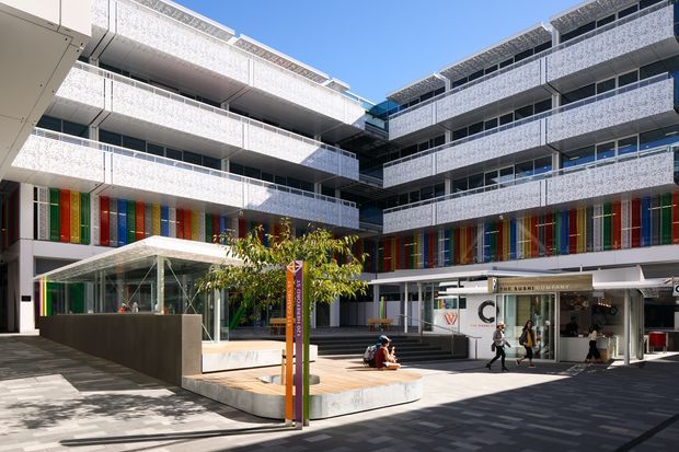 A bright contemporary multi-storey commercial courtyard framed by white patterned balcony bands and colourful vertical facade panels. The open paved forecourt includes integrated seating, a small tree, a glazed pavilion or entrance structure, and active retail or hospitality tenancies at ground level.