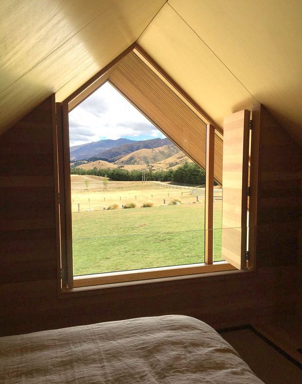 A cozy timber-lined bedroom beneath a sloping roof, featuring a large triangular picture window with timber framing and an angled timber venetian blind. The window opens onto a broad rural landscape of grassland, trees, and rolling mountains. A bed with light neutral bedding occupies the foreground.